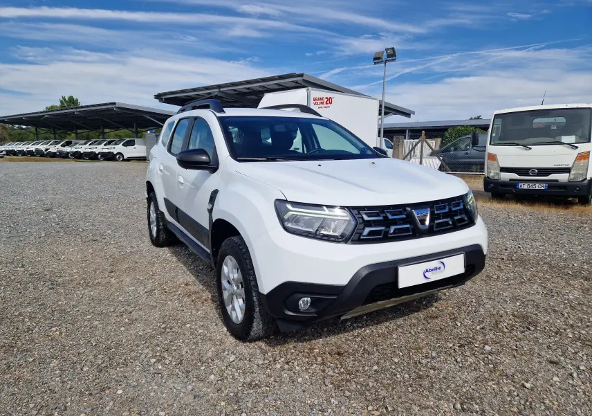 Dacia Duster 4x4 blanc vue 3/4 avant droit sur parking gravillonné avec calandre noire et jantes argentées.