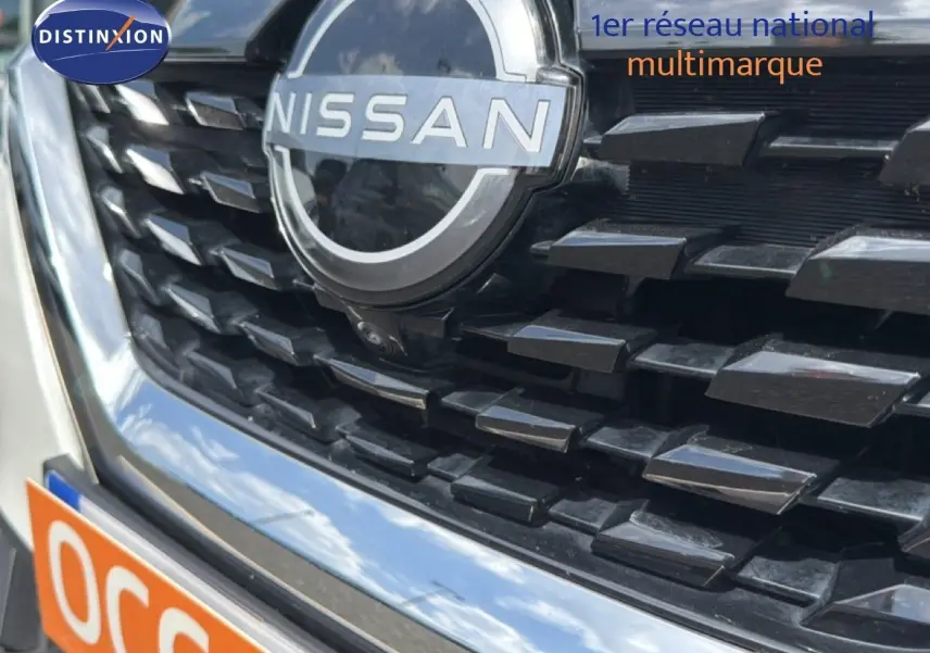 Gros plan sur la calandre noire et le logo Nissan d’un Qashqai blanc nacré 2023, avec reflet du ciel visible.