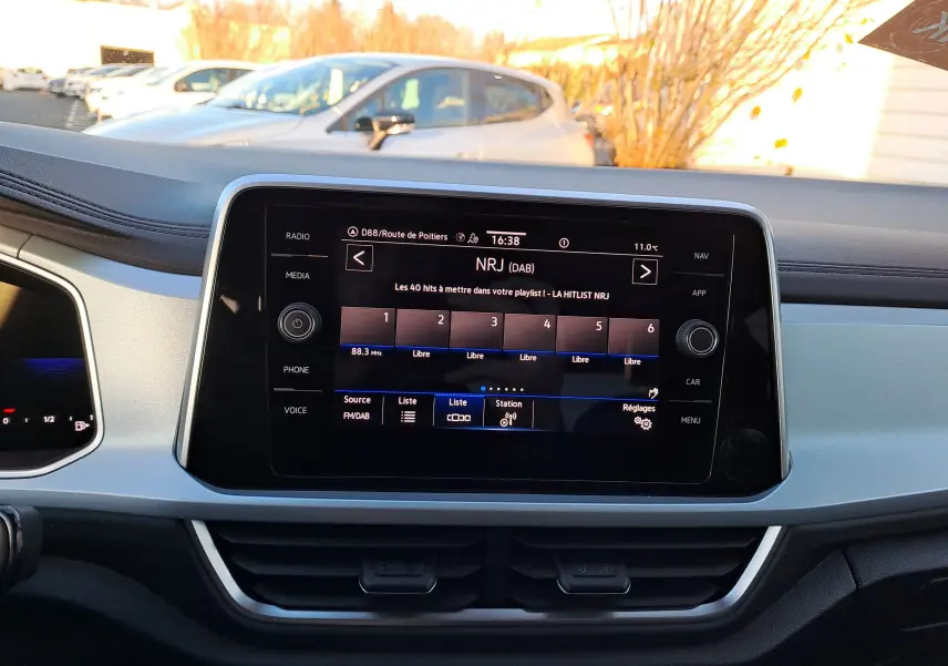 Vue rapprochée de l’écran tactile central du Volkswagen T-Roc gris Ascot, affichant la radio NRJ en extérieur lumineux.