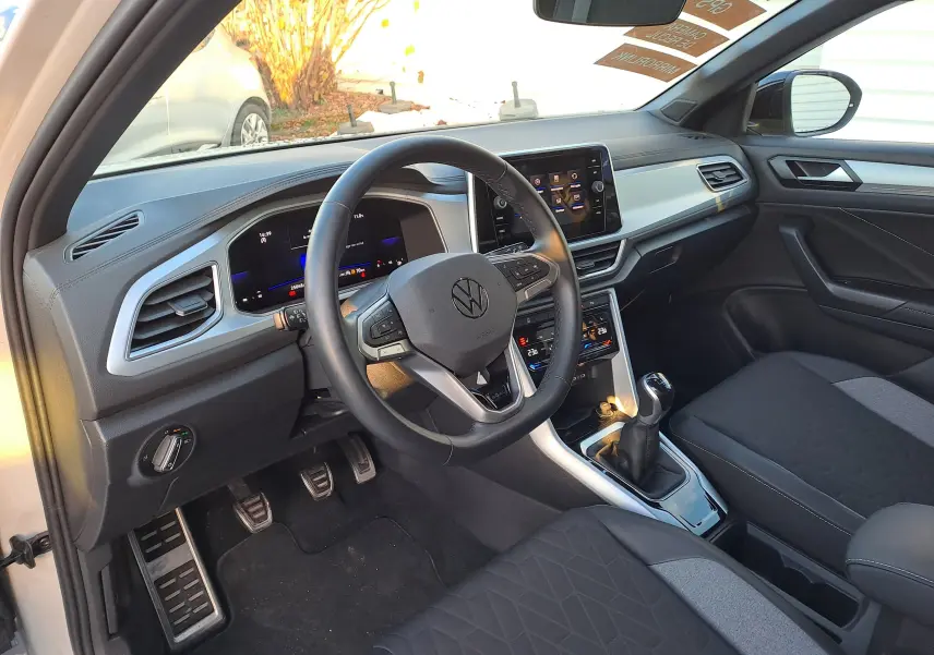 Vue intérieure côté conducteur du Volkswagen T-Roc 2025 gris Ascot, avec volant multifonction et boîte manuelle visible.
