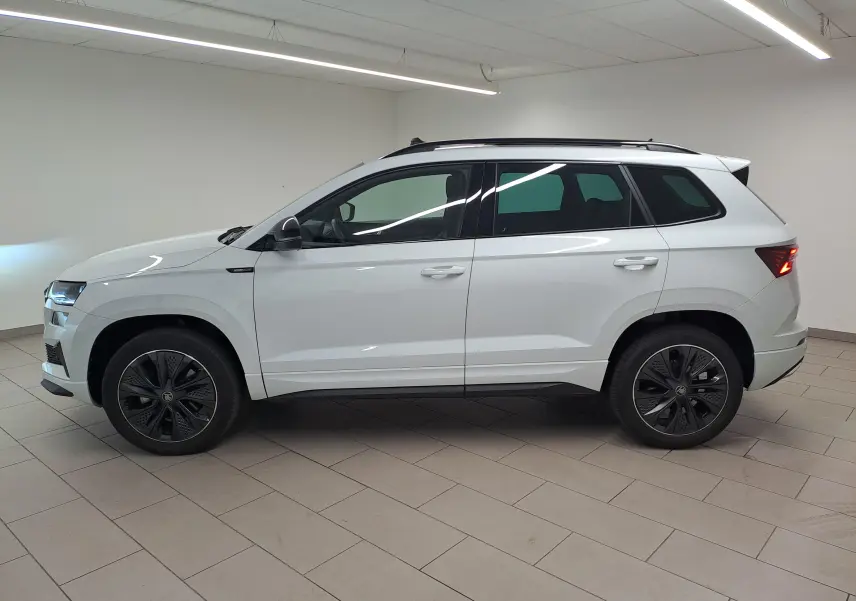 Vue de profil côté gauche d'un Skoda Karoq blanc lune 2025 avec jantes noires et toit ouvrant dans un showroom.