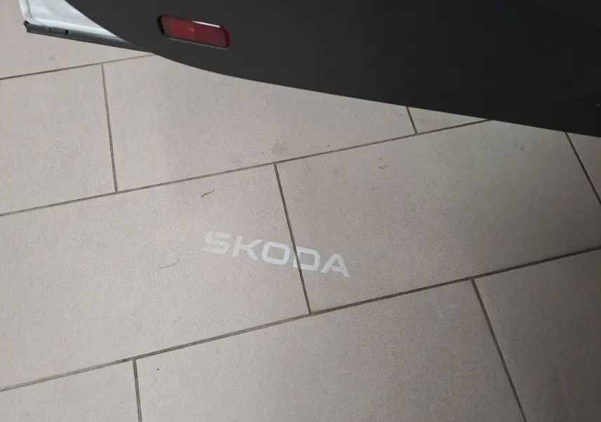 Projection lumineuse blanche du logo SKODA au sol, vue partielle de la porte ouverte d'un SKODA KAROQ Blanc Lune.
