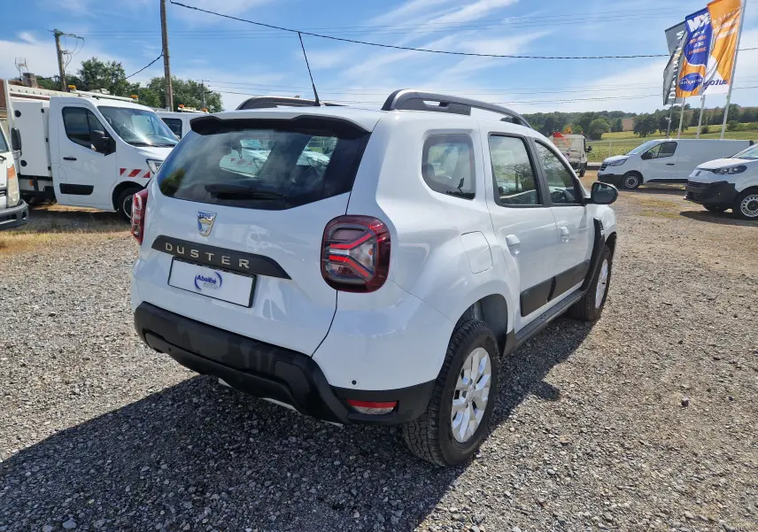 Vue 3/4 arrière droite d'un Dacia Duster blanc 4x4 2023 avec barres de toit et jantes alliage.