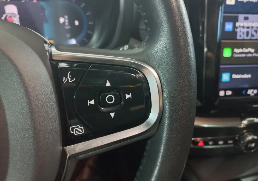 Gros plan sur les commandes vocales et multimédia du volant noir du Volvo XC60 B4 gris clair, avec écran tactile en arrière-plan.
