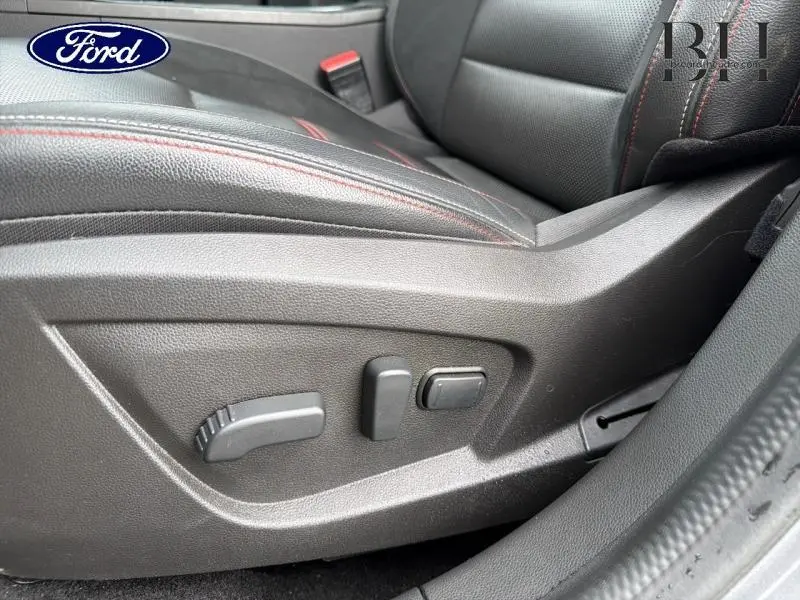 Gros plan sur les commandes électriques du siège avant gauche en cuir noir avec surpiqûres rouges.