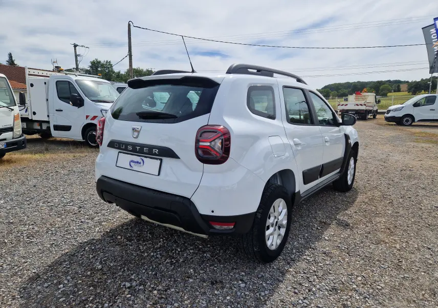 Vue 3/4 arrière droite d'un Dacia Duster blanc 4x4 avec barres de toit et protections noires sur un sol gravillonné.