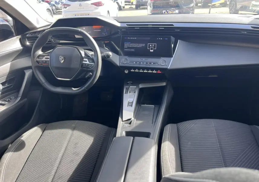 Vue intérieure avant de la Peugeot 308 2024, montrant le volant compact, l'écran tactile central et les sièges tissu gris.