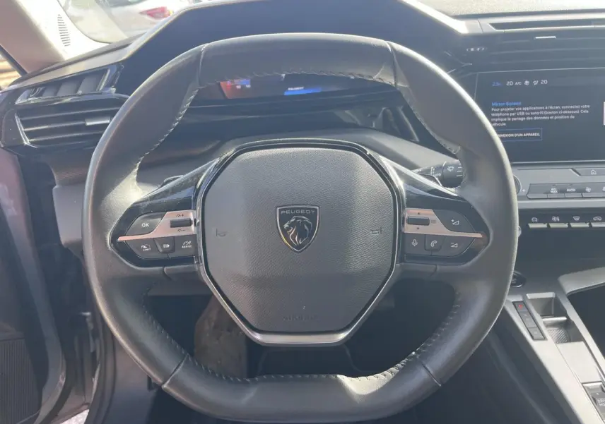 Vue rapprochée du volant multifonction noir avec logo Peugeot, tableau de bord et écran tactile en arrière-plan.