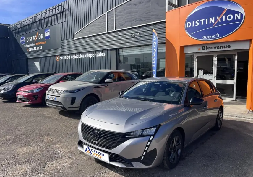 Peugeot 308 gris Artense vue 3/4 avant droit, avec calandre noire et feux LED verticaux, devant concession Distinxion.