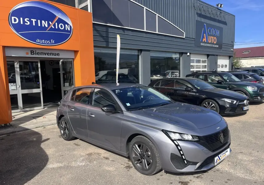 Peugeot 308 gris Artense vue 3/4 avant droit, avec jantes noires et feux LED distinctifs, devant un garage.