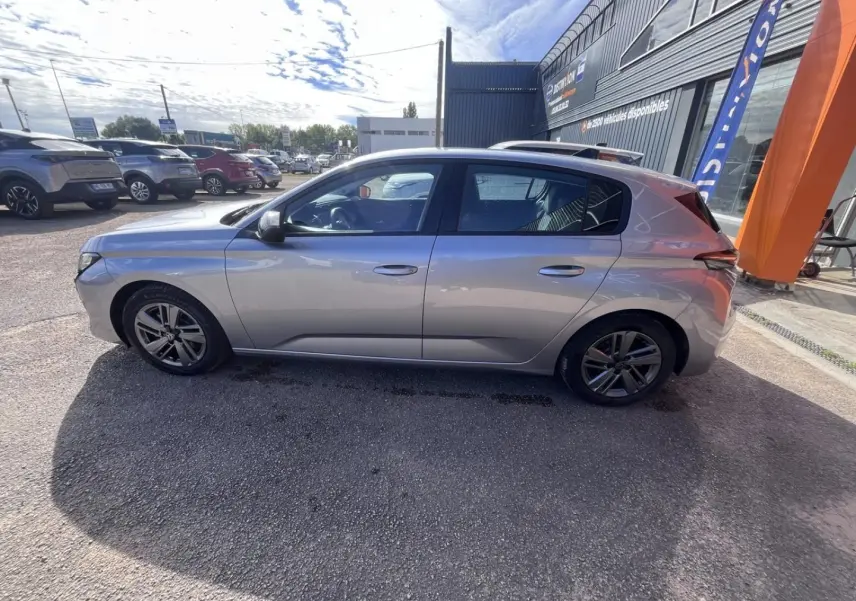 Peugeot 308 gris Artense vue de profil côté gauche, garée devant un bâtiment commercial.