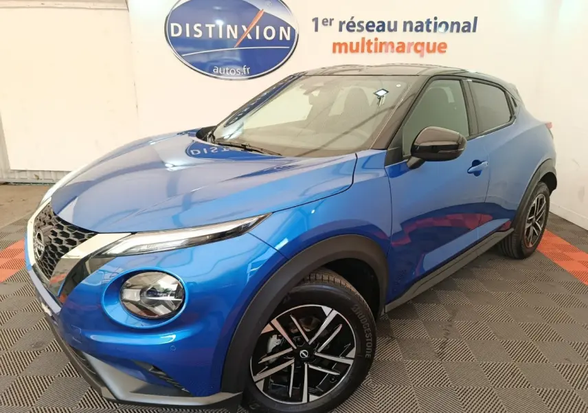 Nissan Juke bleu en 3/4 avant droit, avec toit noir et jantes alliage distinctives, en intérieur showroom.