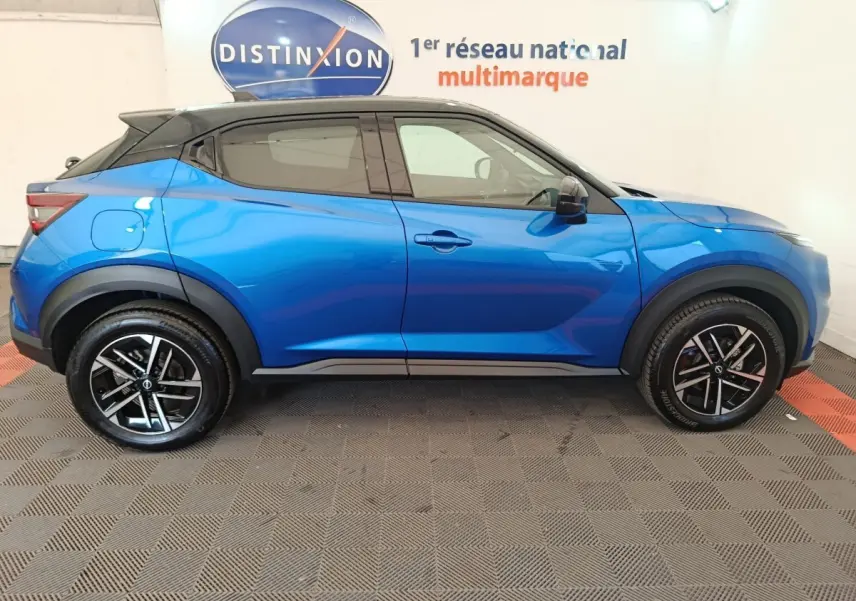 Vue latérale droite d'un Nissan Juke bleu 2025 avec toit noir et jantes alliage noires, en intérieur showroom.