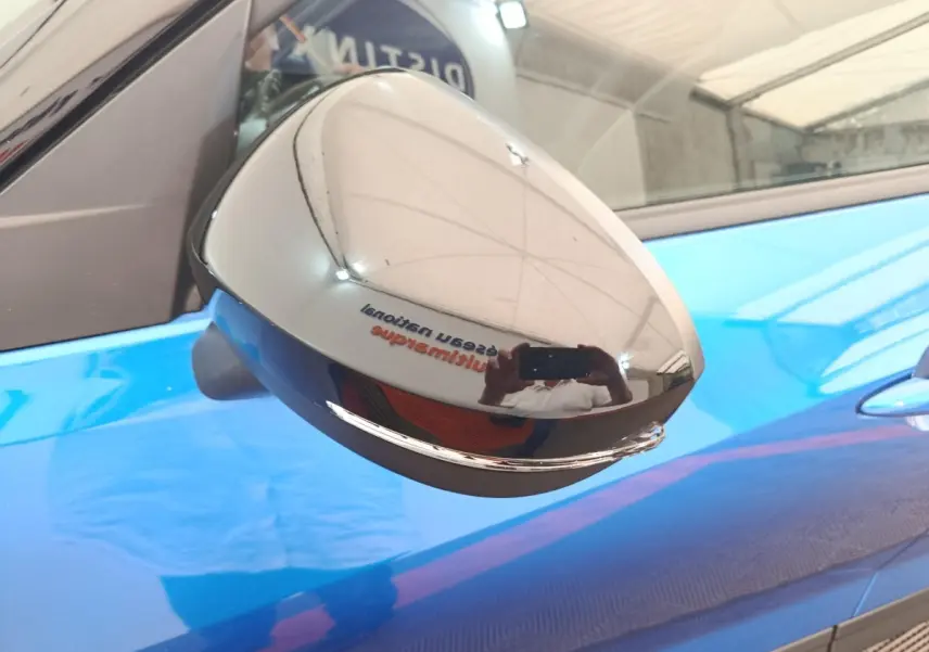 Rétroviseur chromé en gros plan sur le côté gauche d'un Nissan Juke bleu 2025, reflet d'intérieur d'atelier visible.