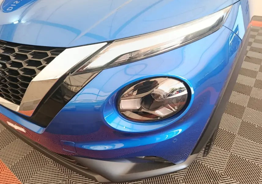 Gros plan sur le phare avant droit bleu vif du Nissan Juke 2025 avec calandre noire et détails chromés.