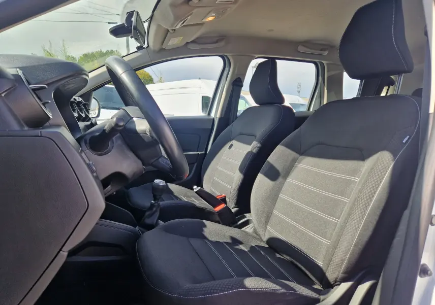 Intérieur du Dacia Duster 2023 blanc, vue côté conducteur montrant les sièges noirs et le volant avec boîte manuelle.