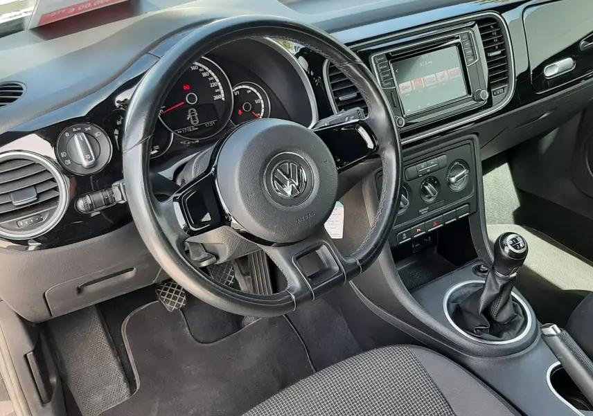Vue intérieure côté conducteur du Volkswagen New Beetle noir, avec volant, tableau de bord et levier de vitesse manuel visibles.