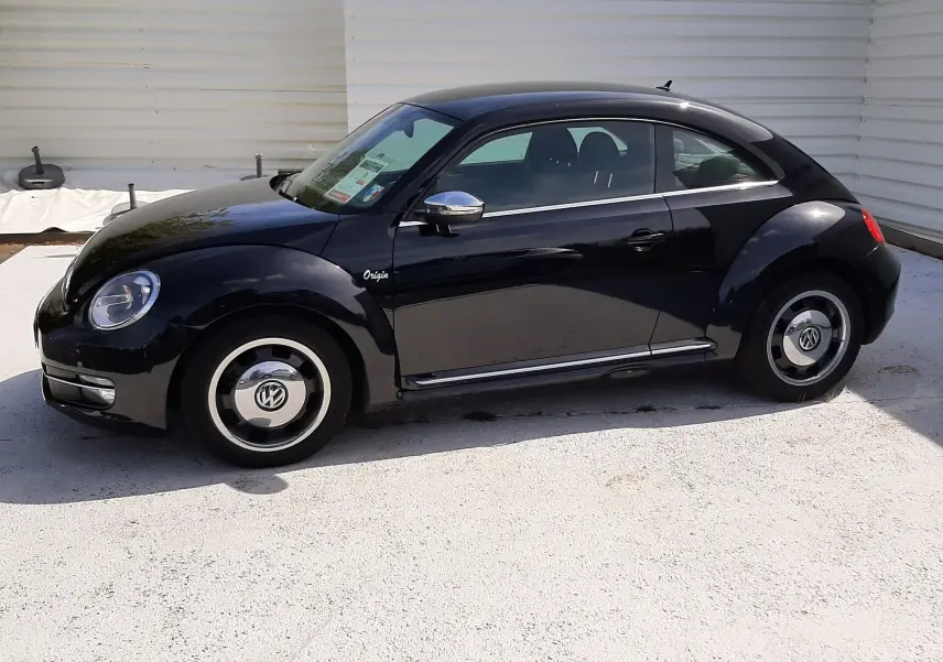 Volkswagen New Beetle noir en vue de profil côté gauche, avec jantes rétro et badge "Origin" visible sur l'aile avant.