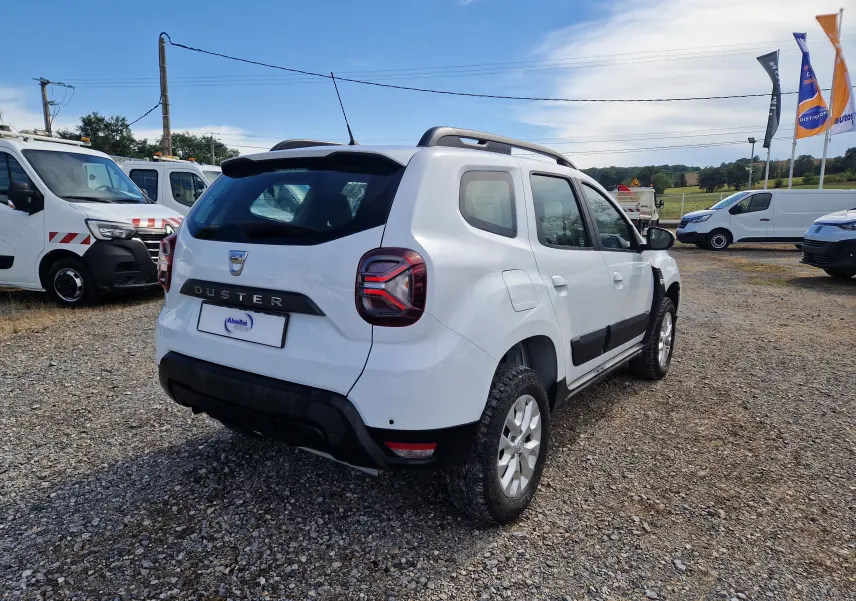 Vue 3/4 arrière droite d'un Dacia Duster blanc 2023 avec barres de toit et protections noires sur un terrain gravillonné.