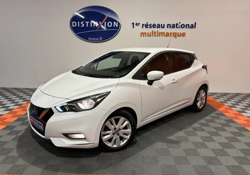 Vue 3/4 avant d'une Nissan Micra blanche 5 portes, en intérieur sur sol gris et orange, avec phares allumés.