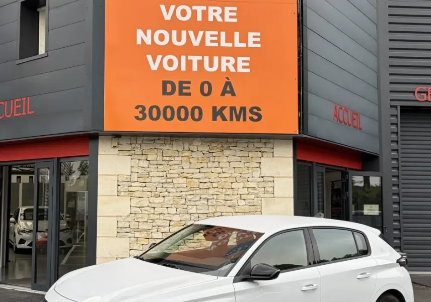 Peugeot 308 blanche vue 3/4 avant droit, avec feux LED verticaux et rétroviseurs noirs devant un bâtiment commercial.