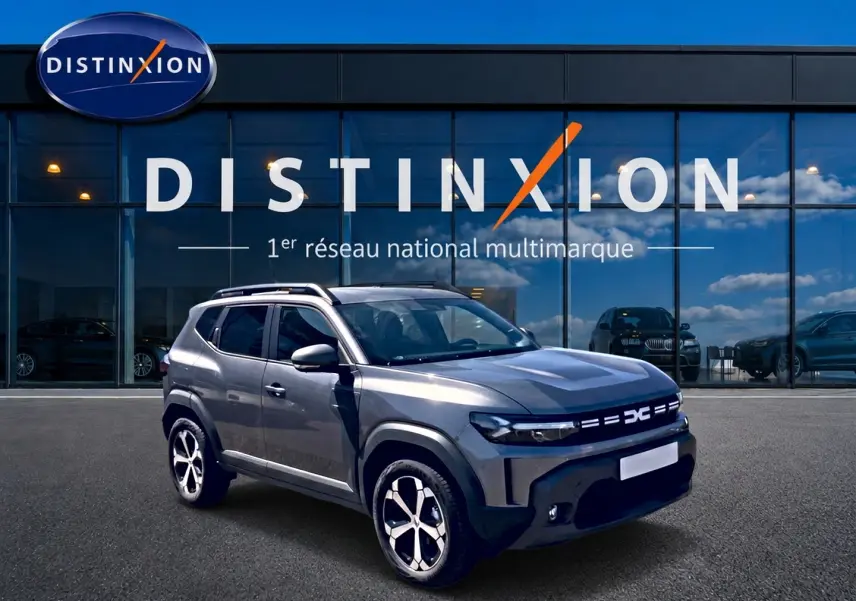 Dacia Duster gris schiste en 3/4 avant droit, avec barres de toit et jantes alu visibles devant un showroom Distinxion.