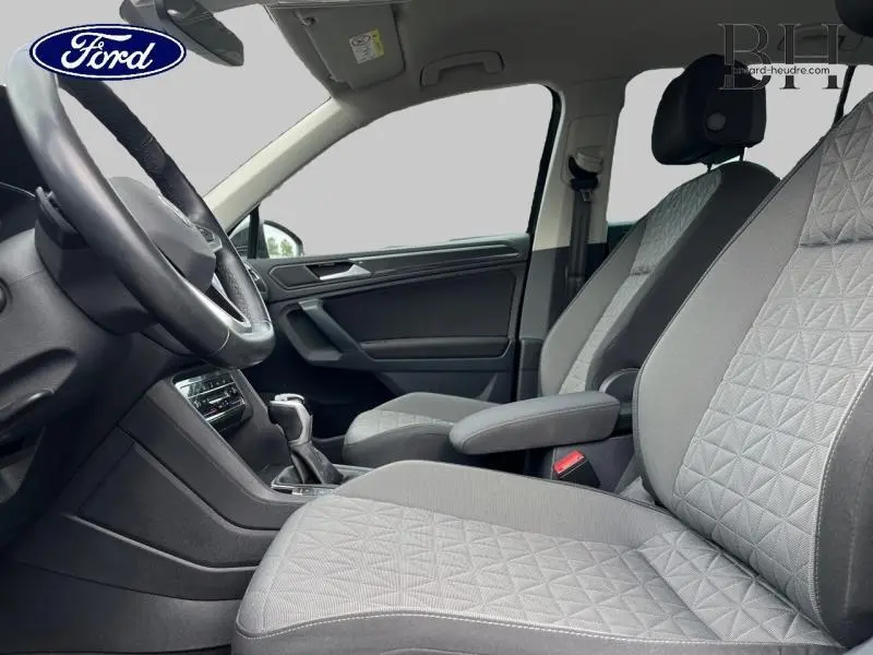 Vue intérieure côté conducteur montrant les sièges tissu gris avec accoudoir central et volant multifonction.