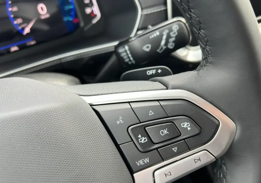 Gros plan sur les commandes multifonctions du volant en cuir du Volkswagen T-Cross gris argent, tableau de bord numérique en arrière-plan.