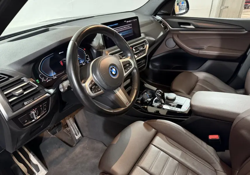 Intérieur BMW X3 xDrive30e 2023 vu côté conducteur, volant cuir, écran tactile et sièges marron cuir.