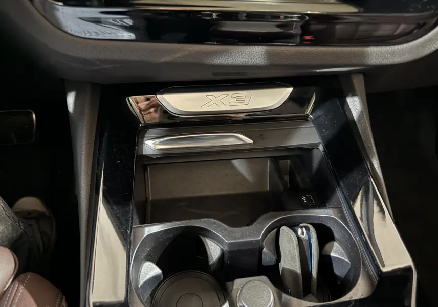 Gros plan intérieur sur la console centrale noire brillante du BMW X3 avec logo X3 et porte-gobelets.