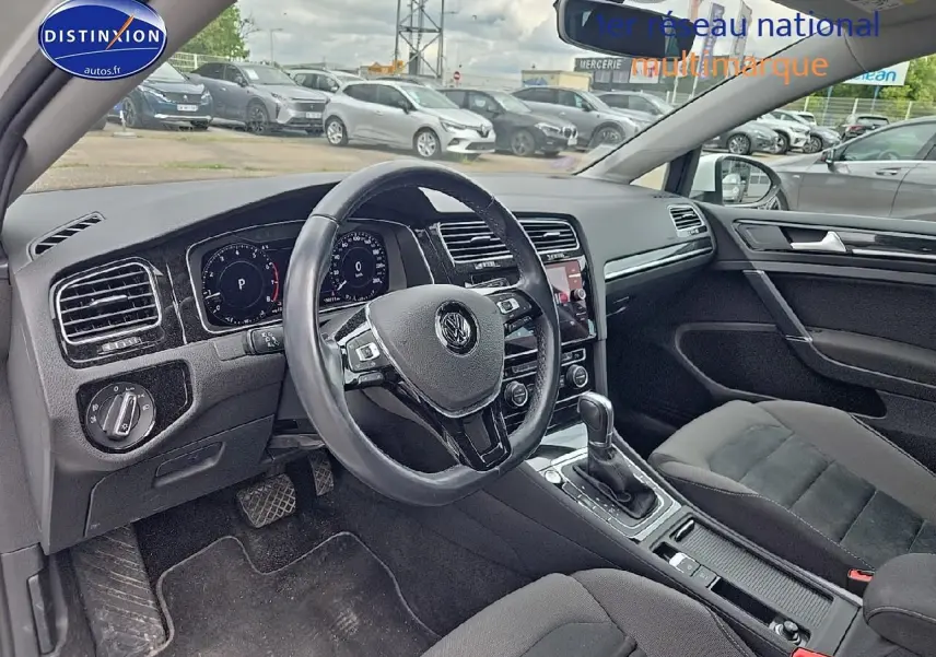 Vue intérieure côté conducteur de la Volkswagen Golf blanche, montrant le volant cuir multifonction et la console centrale.