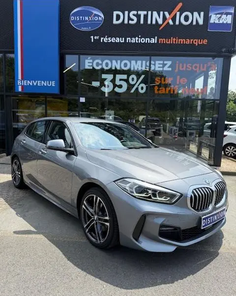 BMW Série 1 118d Edition Sport gris foncé vue 3/4 avant, calandre imposante et jantes alliage visibles devant un showroom.