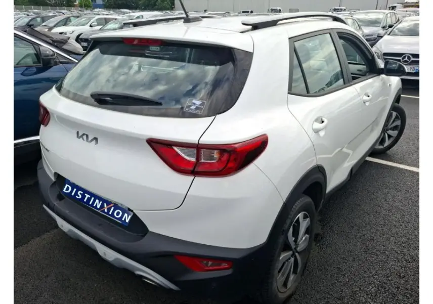 Vue 3/4 arrière droite d’un KIA STONIC blanc 2023 avec feux arrière rouges et toit avec barres de toit noires.