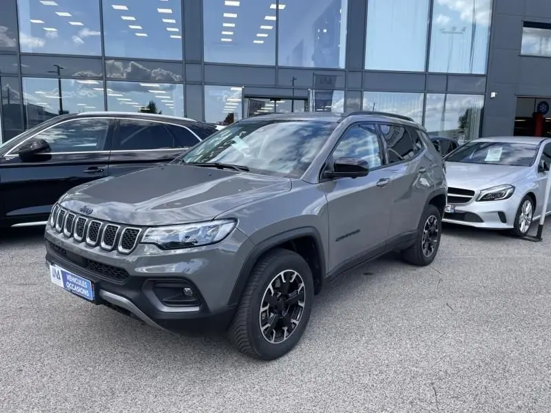 Jeep Compass gris en 3/4 avant droit, avec jantes noires et détails noirs, stationné devant un bâtiment vitré.