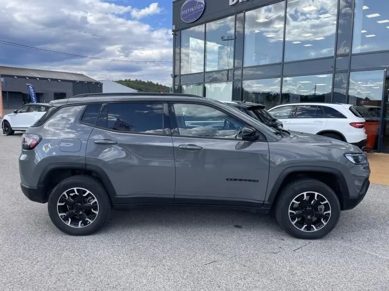 Vue de profil côté gauche d'un Jeep Compass gris 2023 avec jantes noires et vitres teintées, stationné devant un bâtiment vitré.