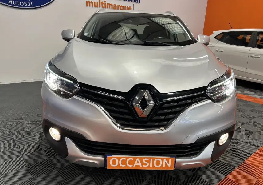 Vue avant d'un Renault Kadjar gris avec phares allumés et plaque "OCCASION" en intérieur showroom.