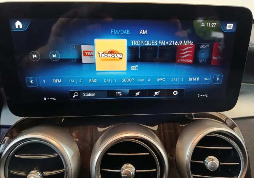 Écran tactile central allumé affichant une radio FM, avec aérateurs ronds et tableau de bord bois dans un Mercedes GLC noir.