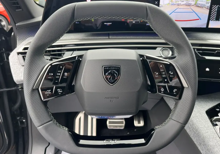 Gros plan sur le volant cuir perforé gris avec logo Peugeot et inscription GT, tableau de bord numérique en arrière-plan.