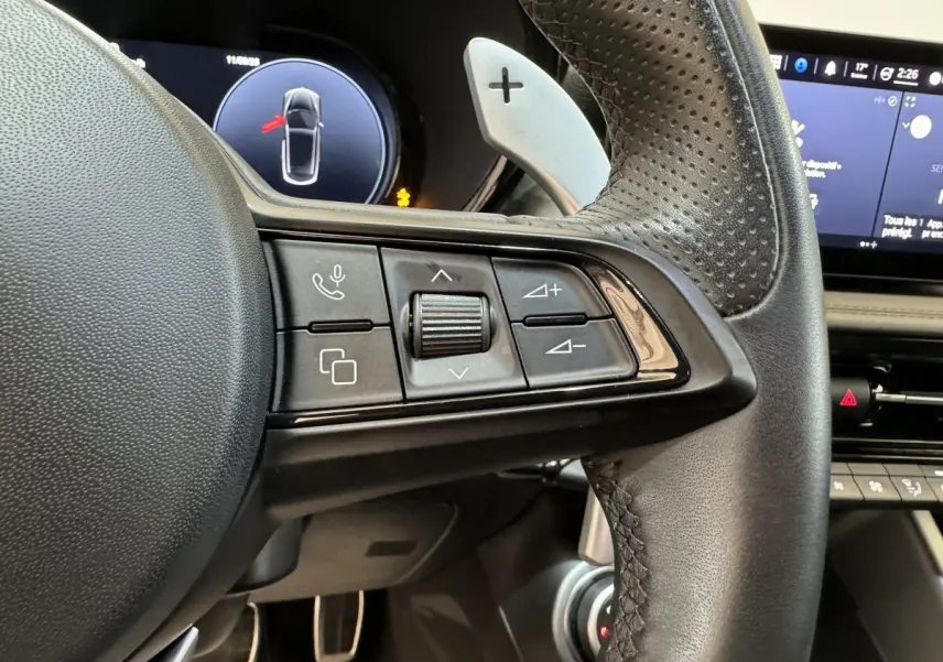 Gros plan sur la partie gauche du volant en cuir perforé de l'Alfa Romeo Tonale rouge avec commandes multifonctions et palettes de vitesse.