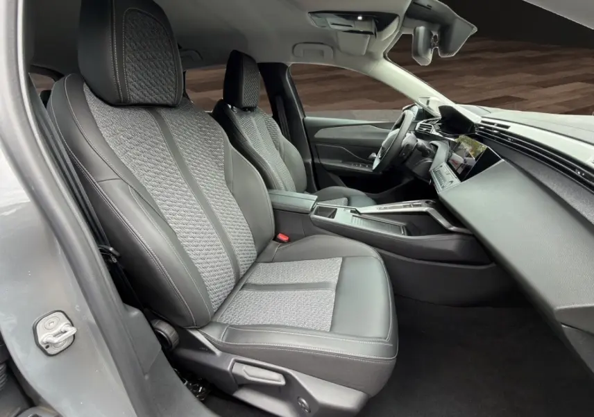 Vue intérieure côté conducteur de la Peugeot 308 Hybrid 145 e-DCS6 Allure 2025 avec sièges gris cuir et tissu, tableau de bord moderne.