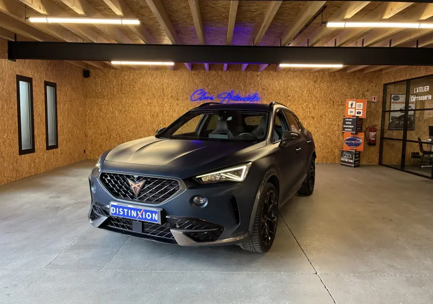 Vue 3/4 avant d'une CUPRA Formentor bleu mat avec jantes noires, dans un showroom au sol gris et mur bois.