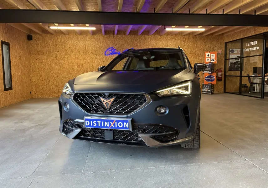 Vue de face d'une CUPRA Formentor bleu mat avec calandre noire et logo cuivre dans un garage en bois.