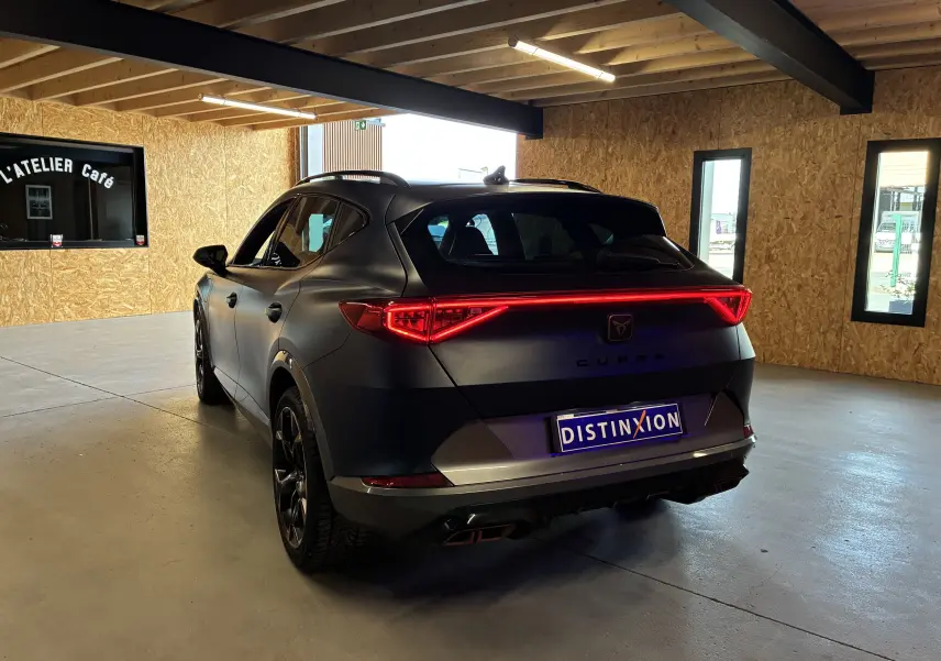 Vue 3/4 arrière d'un CUPRA Formentor bleu mat avec feux arrière LED allumés dans un intérieur en bois clair.