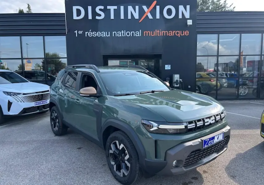 Vue 3/4 avant droit d'un Dacia Duster 2025 kaki dusty avec jantes alliage et barres de toit noires devant un showroom Distinxion.