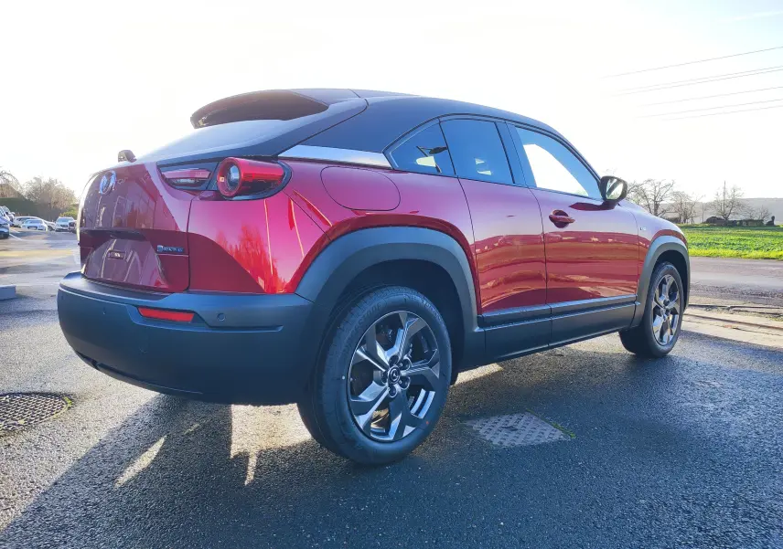 Vue 3/4 arrière droite d'une Mazda MX-30 rouge métal avec toit noir et protections noires sur les passages de roues.