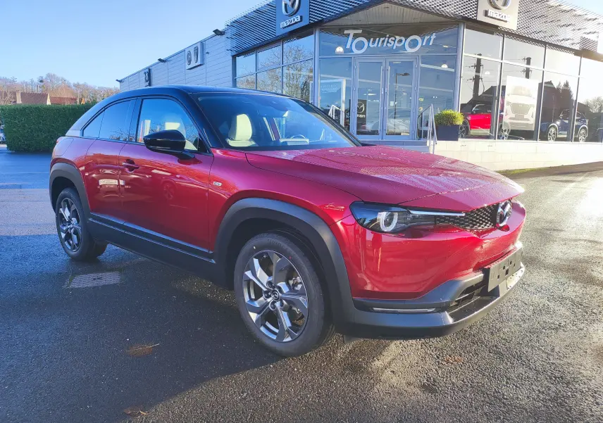 Vue 3/4 avant d'un Mazda MX-30 rouge métal stationné devant un showroom Mazda par temps ensoleillé.