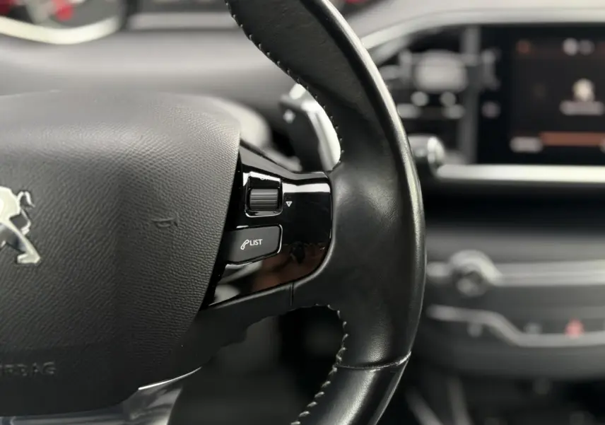 Gros plan sur le volant cuir noir avec commandes intégrées de la Peugeot 308 Allure, tableau de bord flou en arrière-plan.