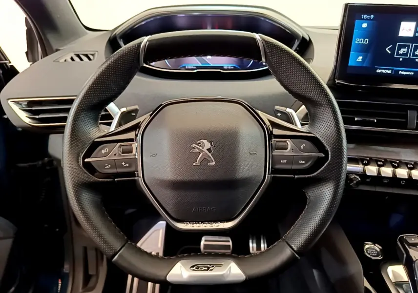 Vue rapprochée du volant cuir noir avec logo Peugeot et badge GT du SUV 5008 bleu, tableau de bord numérique visible.