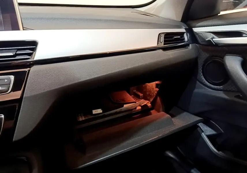 Gros plan sur la boîte à gants ouverte côté passager du BMW X1 noir, avec éclairage intérieur orange et tableau de bord noir.