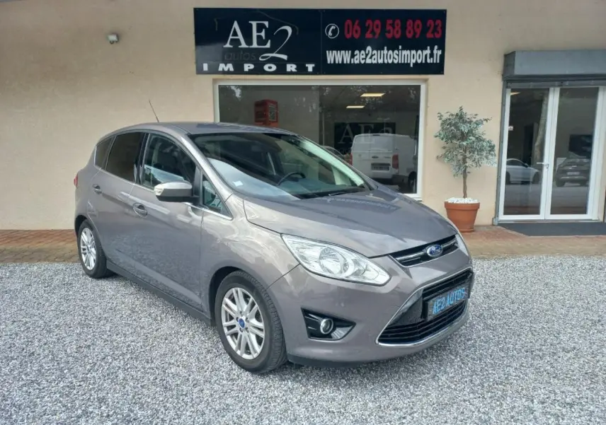 Ford C-MAX gris en 3/4 avant droit, avec calandre chromée et jantes alliage, stationné devant un commerce.